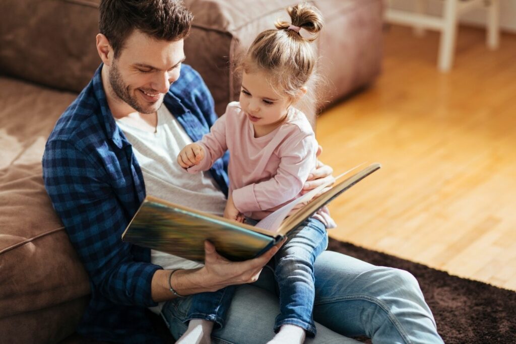 Padre leyendo un libro infantil con su hija pequeña en casa en un momento de lectura compartida