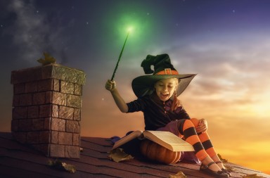 Happy Halloween! Cute cheerful little witch with a magic wand and book of spells. Beautiful child girl in witch costume sitting on the roof, conjuring and laughing.