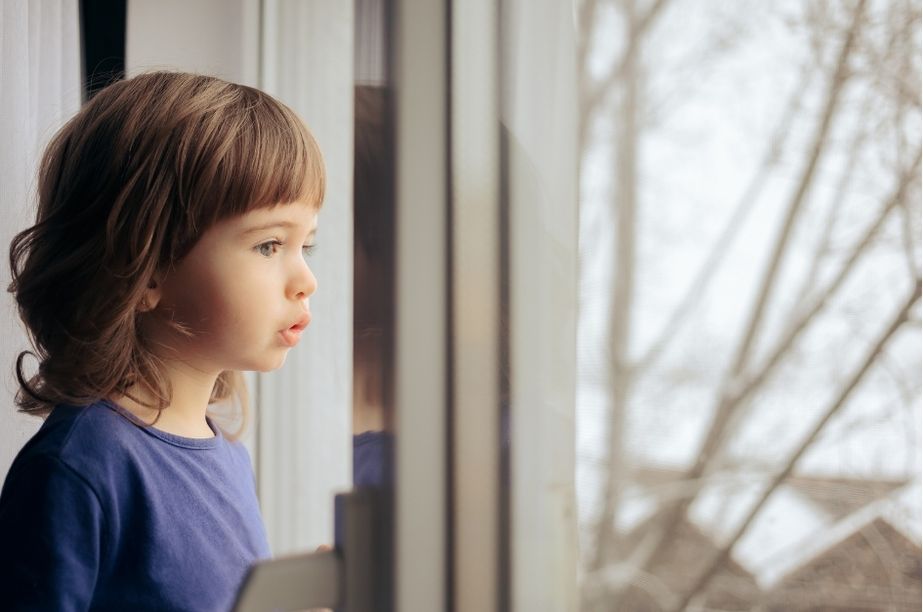 Niña pequeña observando por la ventana con gesto pensativo